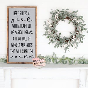 May include: A wooden sign with the text "Here sleeps a girl with a head full of magical dreams, a heart full of wonder and hands that will shape the world." The sign is on a white mantel with a green garland and a wreath.