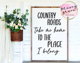 May include: A white wooden sign with black text that reads "COUNTRY ROADS Take me home TO THE PLACE I belong". The sign is hanging on a white brick wall with a small bouquet of pink flowers in a wicker basket to the left.
