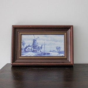 May include: A framed blue and white Delft tile depicting a windmill and a canal scene. The tile is set in a dark wood frame with a gold trim.