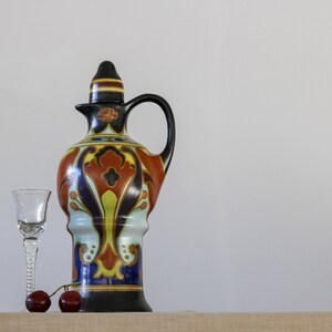May include: A ceramic pitcher with a black, blue, red, and yellow floral design. The pitcher has a spout and a handle. There is a small glass on the left and two red cherries in front of the pitcher.