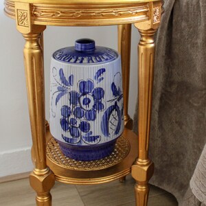 May include: A gold metal stand with a round top and three legs. A blue and white ceramic jar with a floral design sits on the top shelf. The jar has a blue lid.