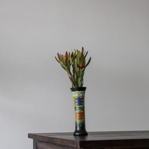 May include: A vase with a colorful geometric pattern, filled with green and orange flowers. The vase is sitting on a dark wooden table.