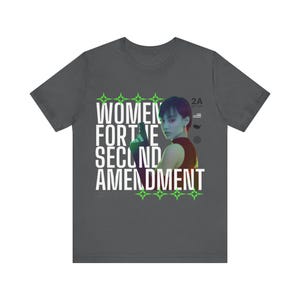 Può includere: T-shirt grigio scuro con la scritta "WOMEN FOR THE SECOND AMENDMENT" in bianco, accentuata da stelle verdi. La maglietta presenta l'immagine di una persona che tiene una pistola. Il testo "2A" è anch'esso visibile.