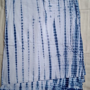 Op de afbeelding: Een witte stof met een blauw tie-dye patroon. De stof is dubbelgevouwen en heeft een verticaal gestreept patroon.