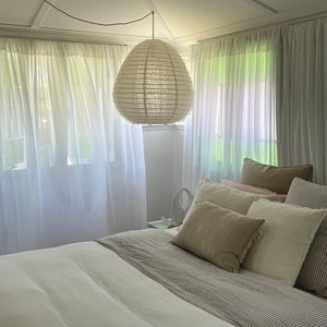 以下が含まれることがあります： 天井から吊り下げられた大きな丸い白い紙製のランプシェードのある白い寝室。ベッドは白い寝具で覆われており、白い枕とベージュの枕がいくつか置かれています。窓には白いシアーカーテンが付いています。