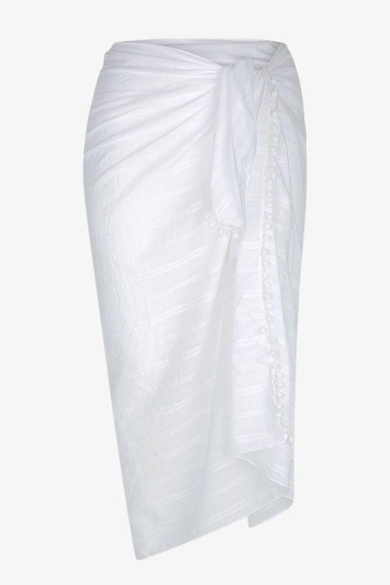 May include: White cotton sarong with a pom-pom trim. The sarong is tied at the waist and has a flowing, relaxed fit.