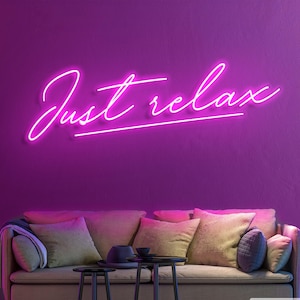 Puede incluir: Un letrero de neón con las palabras "Just relax" en una fuente cursiva, brillando contra una pared púrpura oscura. Debajo, un sofá beige con cojines estampados y pequeñas mesas auxiliares negras crean una escena acogedora.