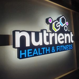 May include: Illuminated sign for "nutrient HEALTH & FITNESS". The sign features the word "nutrient" in white, with "HEALTH & FITNESS" in blue. The logo includes geometric shapes with pink, green, and blue accents, all against a dark background.