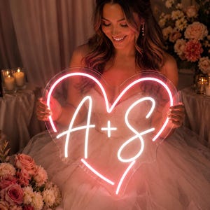 May include: A heart-shaped neon sign featuring the white glowing letters A and S separated by a plus sign. The neon tubing is encased in a clear acrylic backing, emitting a bright pink light that forms the outline of a heart shape.