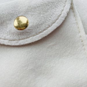 May include: Close-up of a soft, off-white fabric with a rounded flap and a gold-colored button. The fabric has a plush, velvety texture, and the edges are neatly stitched. The button is circular and shiny, contrasting with the matte fabric.