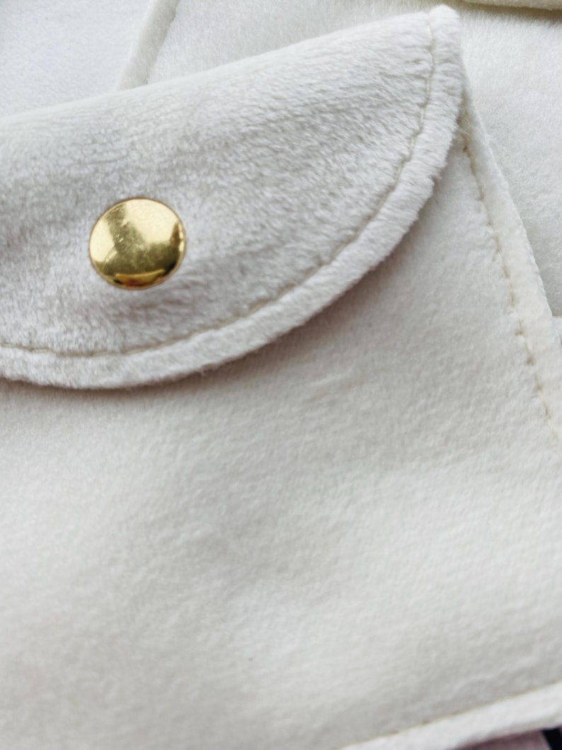 May include: Close-up of a soft, off-white fabric with a rounded flap and a gold-colored button. The fabric has a plush, velvety texture, and the edges are neatly stitched. The button is circular and shiny, contrasting with the matte fabric.