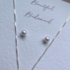May include: A silver necklace and matching pearl stud earrings are displayed on a white card. The card has the words "Beautiful Bridesmaid" written in elegant script. The jewelry is delicate and suitable for a special occasion.