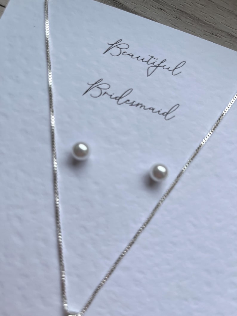 May include: A silver necklace and matching pearl stud earrings are displayed on a white card. The card has the words "Beautiful Bridesmaid" written in elegant script. The jewelry is delicate and suitable for a special occasion.