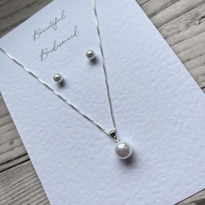 May include: A silver necklace with a single pearl pendant and matching pearl stud earrings are displayed on a white card. The card has the words "Beautiful Bridesmaid" in cursive script.
