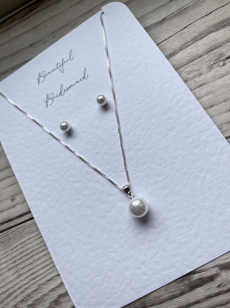 May include: A silver necklace with a single pearl pendant and matching pearl stud earrings are displayed on a white card. The card has the words "Beautiful Bridesmaid" in cursive script.