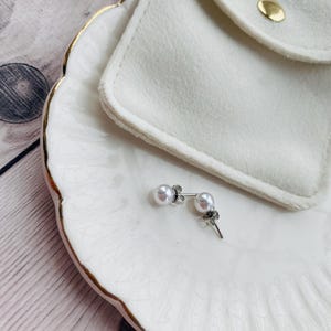 May include: A pair of pearl stud earrings with silver posts and a small, decorative accent. The earrings rest on a white, scalloped-edge dish with gold trim, next to a white pouch with a gold snap closure.