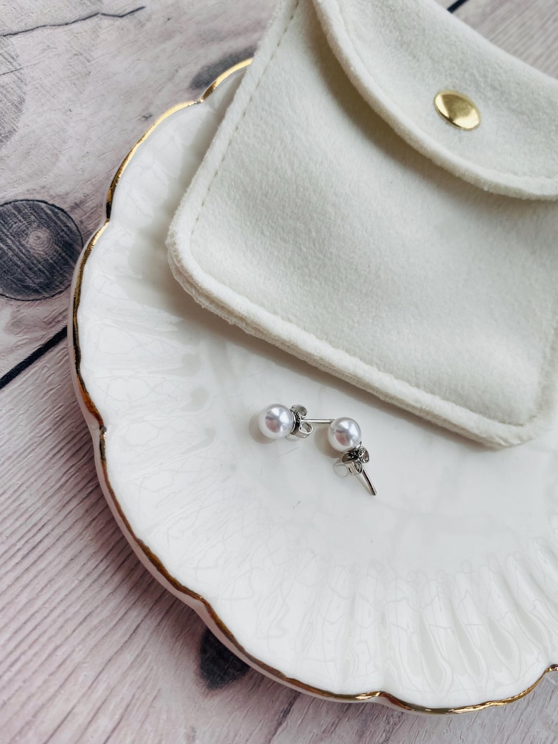 May include: A pair of pearl stud earrings with silver posts and a small, decorative accent. The earrings rest on a white, scalloped-edge dish with gold trim, next to a white pouch with a gold snap closure.