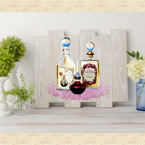May include: Three glass bottles of perfume with decorative labels and stoppers, displayed on a wooden shelf with a pink lace overlay. The bottles are arranged in a row, with the largest bottle in the center. The bottles are surrounded by white flowers and green plants.