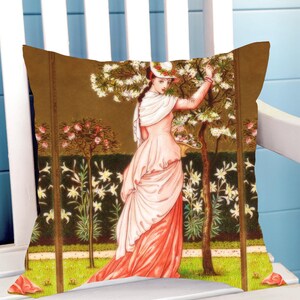 May include: Cushion with a vintage illustration of a woman in a pink dress in a garden. The cushion has a brown background with trees and flowers. The woman is reaching up to pick flowers.