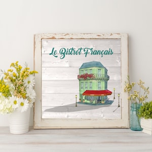 May include: A white framed print with a woodgrain background featuring a cartoon illustration of a green French bistro building with a red awning. The text "Le Bistrot Fran&ccedil;ais" is written in teal above the building.