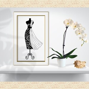 Op de afbeelding: Een zwart-wit illustratie van een paspop met een patroonontwerp. Het kunstwerk wordt tentoongesteld op een witte plank, naast een orchidee in pot en een zeeschelp. De lijst heeft een gouden rand.