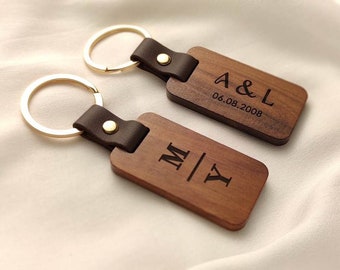 Op de afbeelding: Twee gepersonaliseerde houten sleutelhangers met gouden sleutelringen. De sleutelhangers hebben een rechthoekige vorm met donkerbruine leren bandjes en gouden klinknagels. Eén sleutelhanger is gegraveerd met "A & L" en de datum "06.08.2008". De andere is gegraveerd met "M | Y".