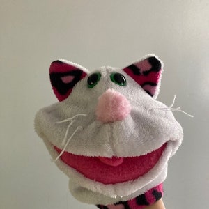 May include: A white plush cat hand puppet with pink and black accents. The puppet has green eyes, a pink nose, and a wide pink mouth. The ears are pink and black. The wrist cuff is pink and black.