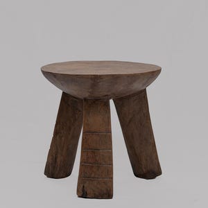 Puede incluir: Un taburete pequeño de tres patas tallado en una sola pieza de madera de color marrón oscuro. El asiento es circular con una superficie ligeramente cóncava, y las tres patas gruesas y robustas presentan líneas horizontales talladas.