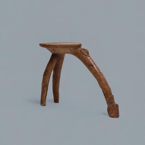 May include: A small, handcrafted wooden stool with a round seat and three legs. The legs are curved and appear to be made from branches, giving it a rustic, natural aesthetic. The stool is a warm brown colour.