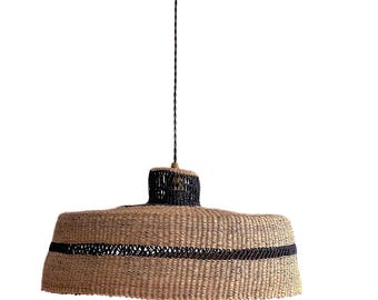 Pode incluir: Uma luminária pendente tecida com um abajur largo, redondo e castanho com guarnição preta. O abajur é feito de materiais naturais e tem um aspeto rústico e artesanal.