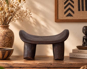 May include: A dark brown wooden headrest featuring a curved, concave top surface and four sturdy, cylindrical legs. The object is carved from a single piece of wood with a smooth, polished finish, showcasing natural wood grain and dark tones.