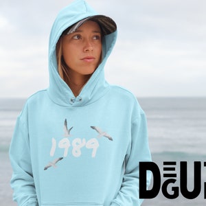 May include: Light blue hoodie with the year "1989" in white script and three seagulls in flight. The hoodie has a drawstring hood and a kangaroo pocket. The person in the image is wearing a camouflage baseball cap.