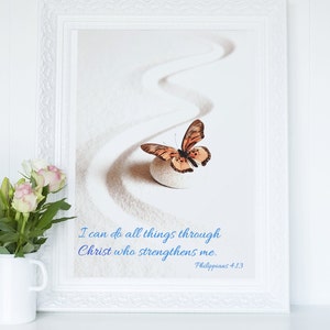 Puede incluir: Una impresión enmarcada en blanco con una mariposa sobre una piedra blanca en un jardín de arena. La impresión contiene la cita "Todo lo puedo en Cristo que me fortalece. Filipenses 4:13"