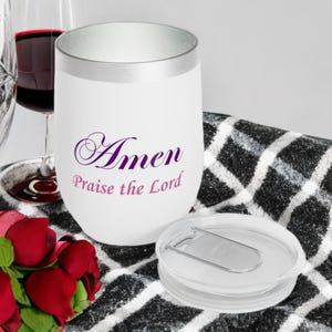 May include: White insulated wine tumbler with a silver rim and a clear lid. The tumbler has the words "Amen Praise the Lord" in purple script. A glass of red wine and roses are in the background.