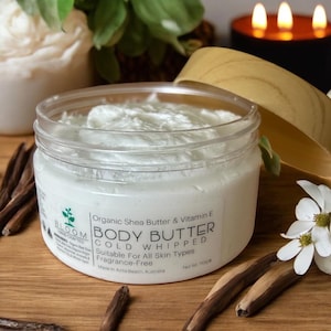 May include: A jar of cold whipped body butter with a white, creamy texture. The label reads "Organic Shea Butter & Vitamin E BODY BUTTER" and "Suitable For All Skin Types". The jar is surrounded by decorative elements including a candle and flowers.