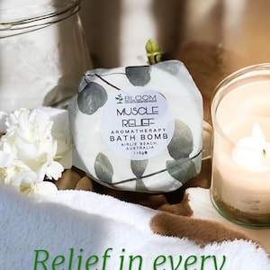 Könnte beinhalten: Aromatherapie-Badebombe mit dem Text "Muscle Relief" und dem "Bloom"-Logo. Die Badebombe ist in Papier mit einem Blättermuster verpackt. Das Bild enthält auch eine brennende Kerze, weiße Blumen und den Text "Relief in every soak".