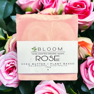 May include: A rectangular bar of pink soap wrapped in a white paper label. The label features the text BLOOM, HAND CRAFTED ORGANIC SOAPS, ROSE, SHEA BUTTER | PLANT BASED, MELBOURNE, AUSTRALIA, and 100g. The soap is surrounded by pink roses.
