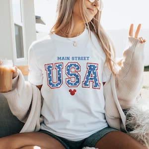 K&ouml;nnte beinhalten: Wei&szlig;es T-Shirt mit einer blauen und roten "USA"-Grafik mit wei&szlig;en Sternen. Die Grafik enth&auml;lt auch den Text "Main Street" und einen roten Mickey Mouse Kopf.