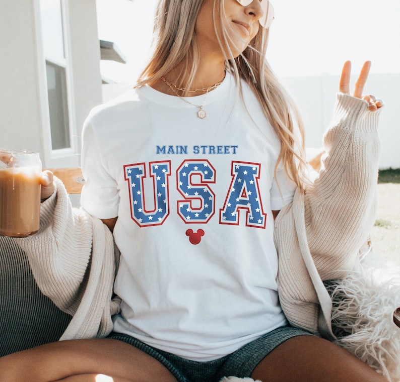 K&ouml;nnte beinhalten: Wei&szlig;es T-Shirt mit einer blauen und roten "USA"-Grafik mit wei&szlig;en Sternen. Die Grafik enth&auml;lt auch den Text "Main Street" und einen roten Mickey Mouse Kopf.