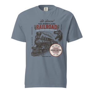 May include: A blue t-shirt featuring a vintage-style graphic of a steam train, with the text "All Aboard! The Disneyland Railroad" and "Don't Miss the Grand Canyon Diorama." The design includes a dinosaur silhouette.