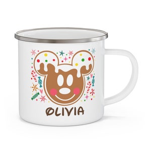 May include: White enamel mug with a gingerbread Mickey Mouse design. The design includes a brown gingerbread Mickey face with white icing and colorful sprinkles, surrounded by candy and the name "OLIVIA".