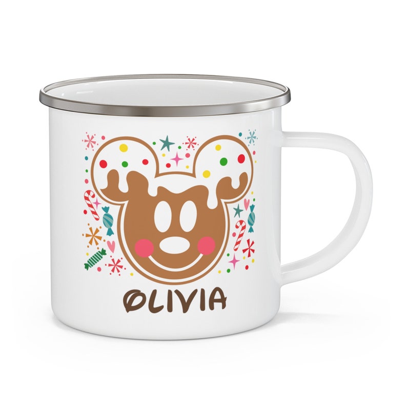 May include: White enamel mug with a gingerbread Mickey Mouse design. The design includes a brown gingerbread Mickey face with white icing and colorful sprinkles, surrounded by candy and the name "OLIVIA".