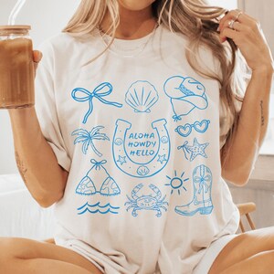 Peut inclure: T-shirt cr&egrave;me avec des illustrations bleues d'un n&oelig;ud, d'un coquillage, d'un chapeau de cowboy, de lunettes de soleil, d'un palmier, d'un fer &agrave; cheval avec du texte, d'un bikini, d'un crabe, d'une &eacute;toile de mer, d'un soleil et d'une botte de cowboy. Le texte sur le fer &agrave; cheval dit "Aloha Howdy Hello".