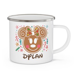 May include: White enamel mug with a gingerbread Minnie Mouse design. The design includes a brown gingerbread face with a bow, pink cheeks, and the name "DYLAN" in brown text. Colorful candy and snowflake accents surround the character.