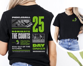 Może przedstawiać: Czarny T-shirt z zielonym i białym nadrukiem graficznym z tekstem "Pickleball", "Dominate the Courts", "25", "Day Dinker" i "Stay Out of the Kitchen". Grafika zawiera również kort do pickleballa z graczami i piłką do pickleballa.