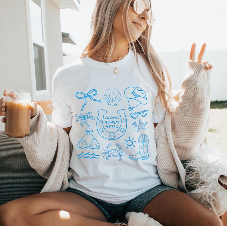 Peut inclure: T-shirt blanc avec des illustrations bleues repr&eacute;sentant un n&oelig;ud, un coquillage, un chapeau de cowboy, des lunettes de soleil, un palmier, un fer &agrave; cheval, un bikini, un crabe, une &eacute;toile de mer, un soleil et une botte de cowboy. Le texte "ALOHA HOWDY HELLO" est &agrave; l'int&eacute;rieur du fer &agrave; cheval.