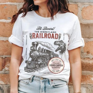 May include: White t-shirt featuring a vintage-style illustration of a steam train, a dinosaur, and the text "All Aboard! The Disneyland Railroad." Includes "Don't Miss the Dramatic Grand Canyon Diorama EST. 1958."