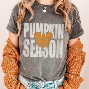 May include: Dark gray t-shirt with the words "PUMPKIN SEASON" in white block letters. An orange pumpkin with mouse ears is in the center. The shirt is worn with a gold-colored sweater.