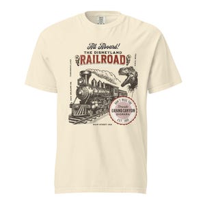 May include: Cream-colored t-shirt featuring a vintage-style illustration of a steam train on tracks, with the text "All Aboard! The Disneyland Railroad." Includes a dinosaur graphic and text that reads "Don't Miss The Grand Canyon Diorama Est. 1958."
