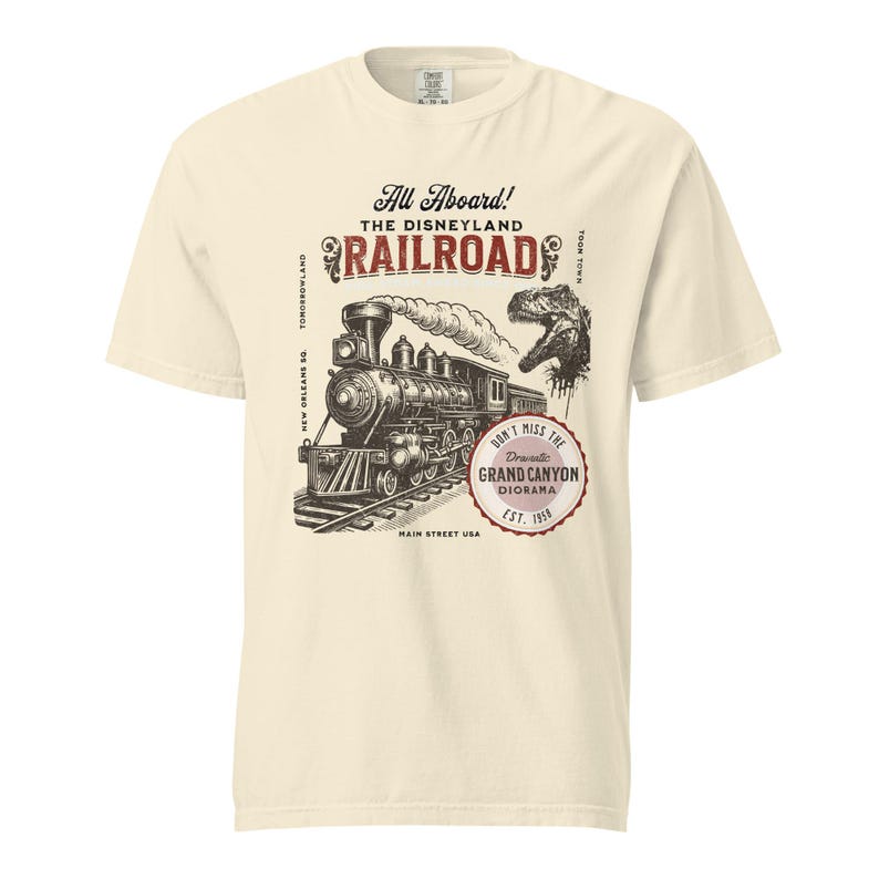 May include: Cream-colored t-shirt featuring a vintage-style illustration of a steam train on tracks, with the text "All Aboard! The Disneyland Railroad." Includes a dinosaur graphic and text that reads "Don't Miss The Grand Canyon Diorama Est. 1958."